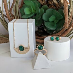 Une présentation avec une plante verte d'un collier , d'une bague et d'une paire de boucles d'oreilles qui sont en plaqué or et malachite, une pierre semi-précieuse verte. Une collection du fabricant bijoutier Thabora.