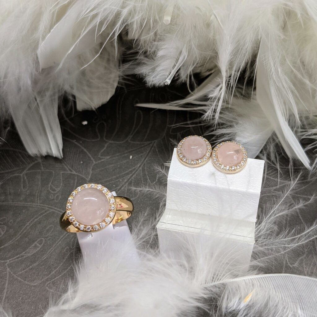 Avec un décor de plumes blanches, présentation d'une bague et d'une paire de boucles d'oreilles de la marque Thabora en plaqué or et quart rose, style cabochon avec contour d'oxydes blancs